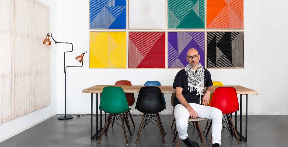 Matthias de Vogel aka artis fault lines lab sits in front of his colourful, primary colour woven contemporary canvas works in square forms next to a contemporary dining table