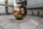 A bronze metal spherical sculpture reflecting the city of Dublin, whilst passerby's walk alongside it.