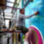 An artist works on his mural, graffiti spraying a pink line over a blue mural on an exterior public art façade, whilst sitting on scaffolding.