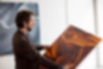 © Artelier Artist Christoph Schrein looking at his abstract, orange canvas painting artwork at an angle with one of his large blue paintings blurred in the background