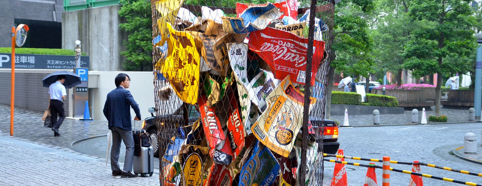 60. © Photo by Nick West showing an above human size garbage bin trash can filled with leading industry brand packaging like coca cola in a japanese urban space, left to the artwork is a business suit man with a suitcase and another business suit man with an umbrella in front of a parking lot and right to the can are 3 orange traffic cones