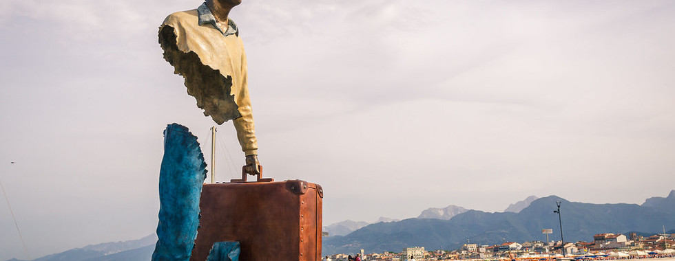18. © Photo by Pat_4light showing a human figure sculpture looking over the water of a port carrying a suitcase but parts of his body are missing such as his torso and upper leg