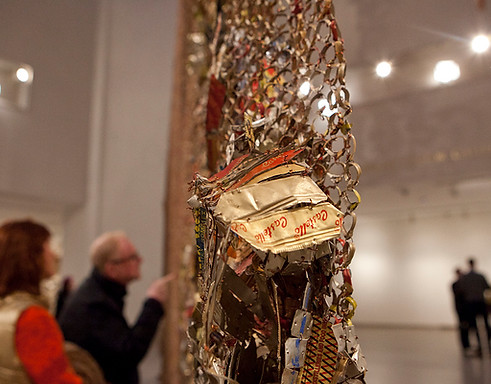 close up detai shot of artist el anatsui's artwork made of threaded recycled metal and wire in hues of yellow also featuring exhibition guests inspecting the artwork up close in the background blurred