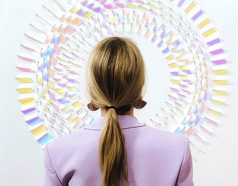 back view of blond woman with pink tuxedo inspecting a chris wood dichroic wall art piece in circular form, artist is managed by artelier art advisory