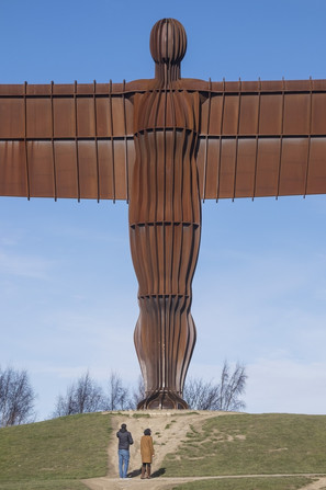 A close-up of angel of the north, figure in bronze with arms cut off and two people viewing it in the front.