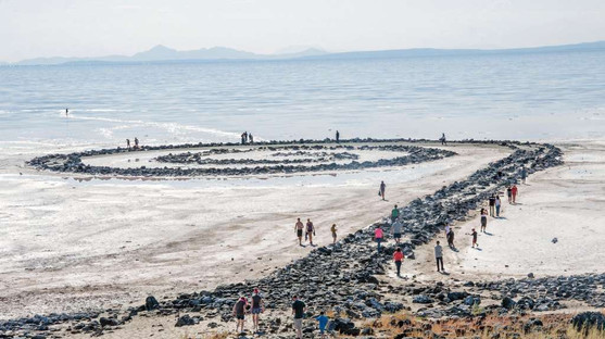 A close-up, panaromic view of Spiral Jetty by Robert Smithson, with people in the foreground surrounding the spiral land art installation on a cloudy day.
