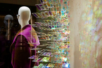 Dynamic glass artwork shown from a side wide-angle lens, with a mannequin to the left. The glass art reflects coloured hues.