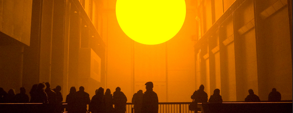 © Photo by Tom Trickland, low glow sun in the tate as a contemporary art installation display with shadowed figures looking up at it in the centre of the space, overall tint of the photo is warm orange yellow