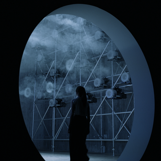 A.A.Murakami’s The Passage of Ra (2024) is a multimedia art installation inspired by Ra, the sun deity in ancient Egyptian mythology, and his daily passage across the sky to create day and night.  The artwork features a screen projection of a digital seascape and tangible fog rings that float toward it. Upon contact with the wall, the rings vanish into the air and appear on screen to sail into the ocean horizon, resuming their journey in a virtual world. Ra’s never-ending voyage is reflected in the fog rings, which transcend our physical space to achieve eternity in the digital realm.  In this liminal space between art and technology, the viewer is invited to draw parallels between the short-lived fog rings and the fleeting nature of our existence, both destined to break down and transform into new states of energy across dimensions.