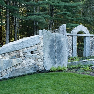 A stone dry wall with an arch at the centre of stacked stone to create an organic contemporary public art piece set against a forest.